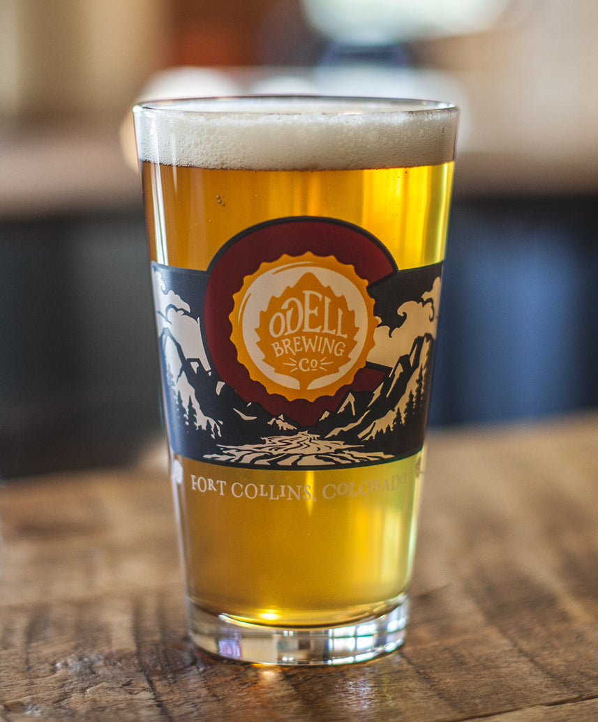 A pint glass full of beer with mountains, a river, and the Odell Brewing logo in the middle of the "C" from the Colorado flag.